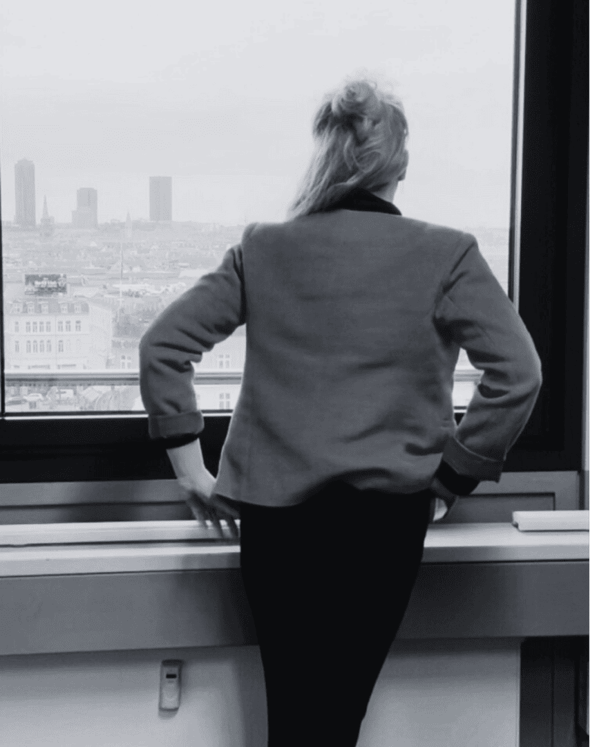 About gyde and patentsmarter™ | patentsmarter,gyde Person with long hair in a jacket standing indoors, looking out a window at a cityscape with tall buildings in the background.
